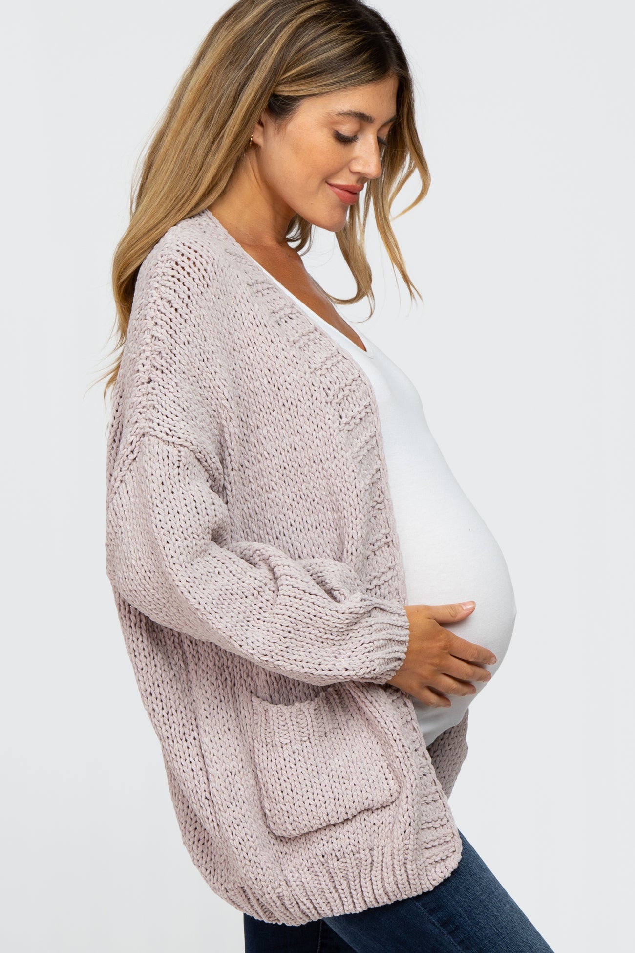 woman wearing a light pink maternity cardigan, holding her baby bump, paired with a white top and dark jeans