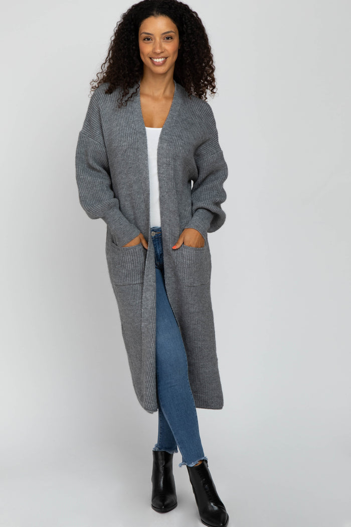 woman wearing a long gray knit cardigan with pockets, white top, and blue jeans, standing and smiling