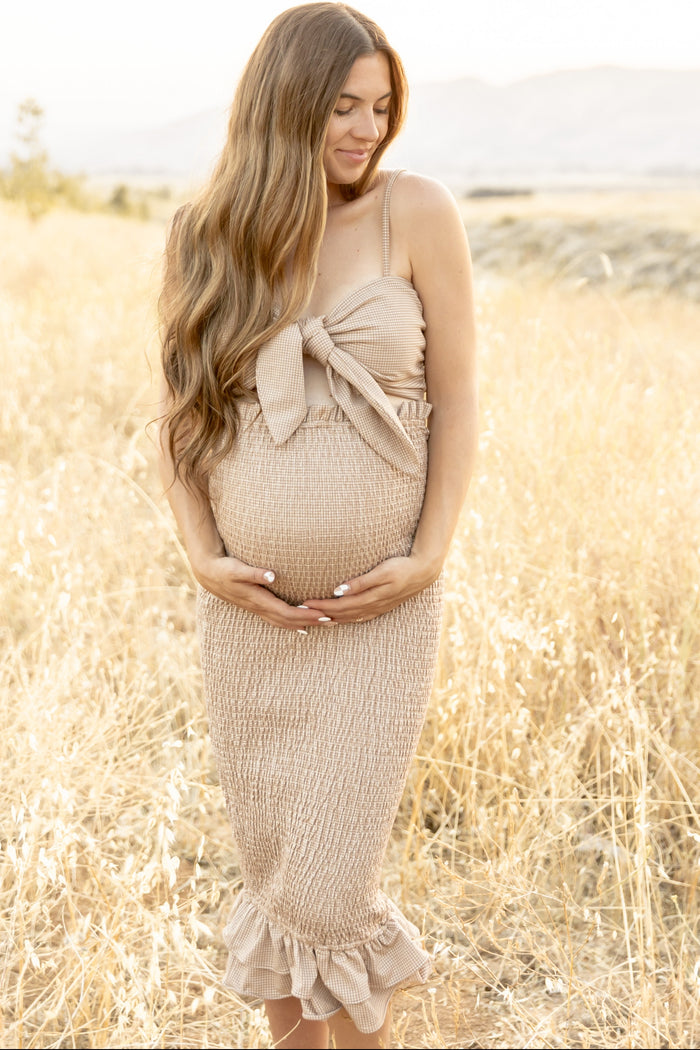 Pregnant woman in a tan gingham dress holding her baby bump in a golden field during a maternity photoshoot