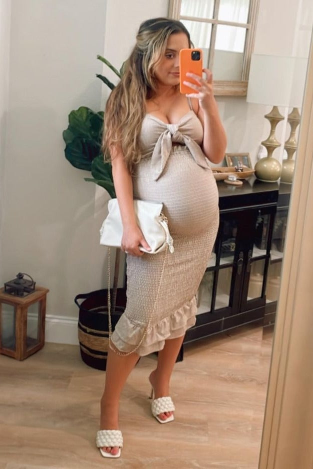 woman wearing a beige maternity dress with a bow detail and ruffle hem, holding a white handbag and showcasing her baby bump