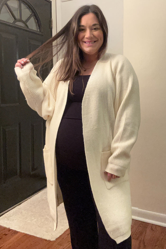 woman wearing a cream long sleeve maternity cardigan over a black dress and displaying her baby bump