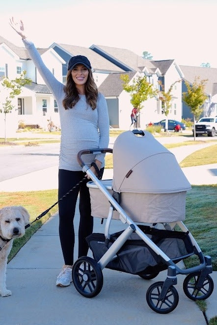 woman wearing a light gray maternity top and black leggings, pushing a beige stroller while walking a dog