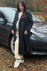 Pregnant woman in a long black coat and cream sweatpants standing in front of a grey Tesla car outdoors