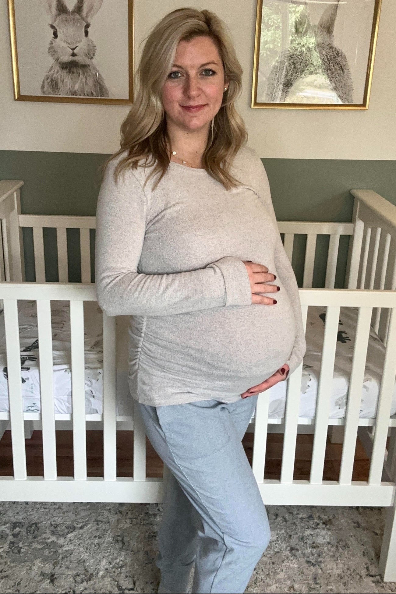 woman wearing a light gray long sleeve maternity top and gray joggers, holding her baby bump in a nursery setting