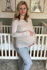 woman wearing a light gray long sleeve maternity top and gray joggers, holding her baby bump in a nursery setting