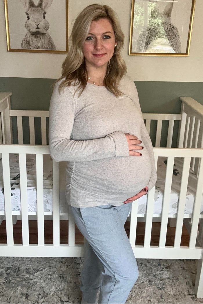 woman wearing a light gray long sleeve maternity top and gray joggers, holding her baby bump in a nursery setting