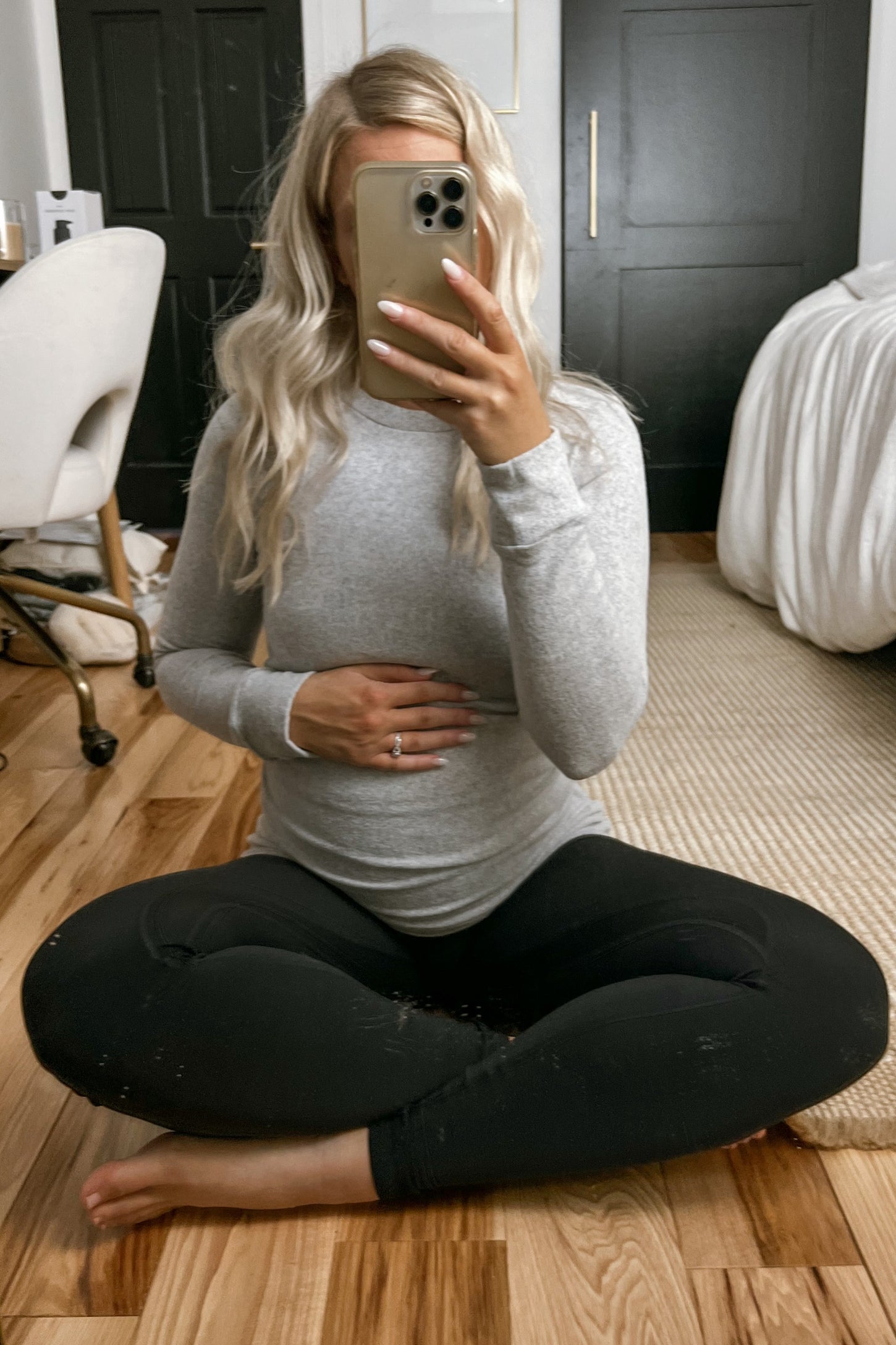 woman wearing a gray long sleeve maternity top and black leggings, holding her baby bump while sitting on the floor
