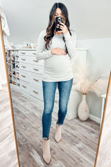 woman wearing a light grey long sleeve maternity top and blue skinny jeans, holding her baby bump in front of a mirror