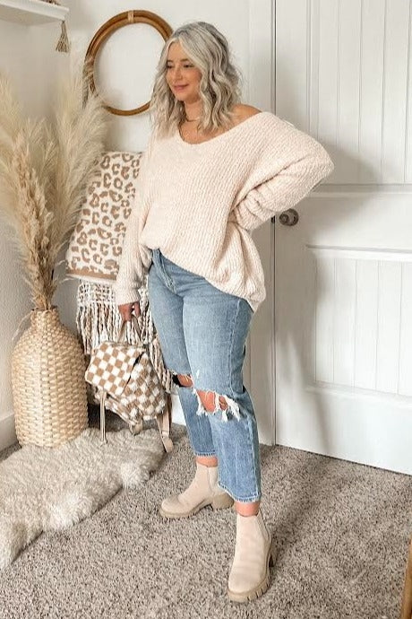 woman wearing a light pink oversized sweater, distressed jeans, and beige ankle boots, standing in a cozy setting
