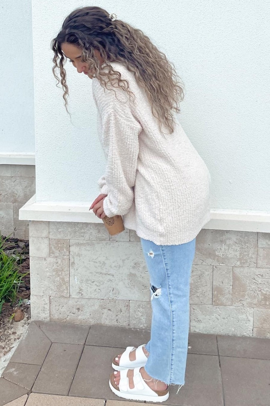 woman with long curly hair wearing cream fuzzy sweater, distressed light wash jeans, and white sandals
