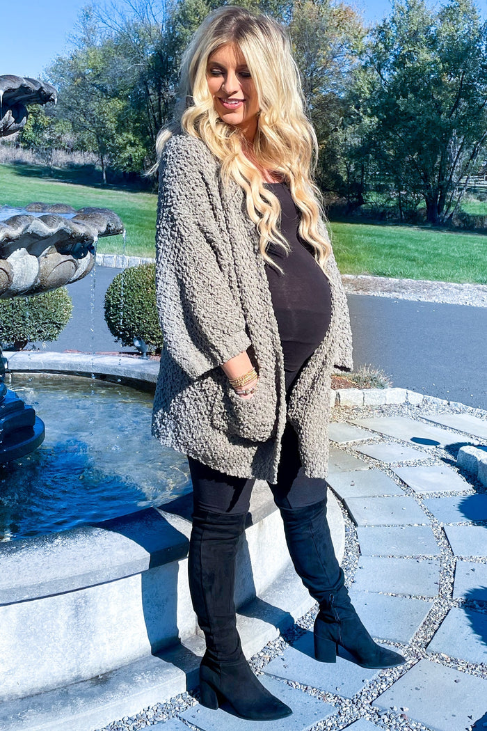 woman wearing a dark brown maternity top, gray cardigan, black leggings, and black heeled boots, showing her baby bump outdoors