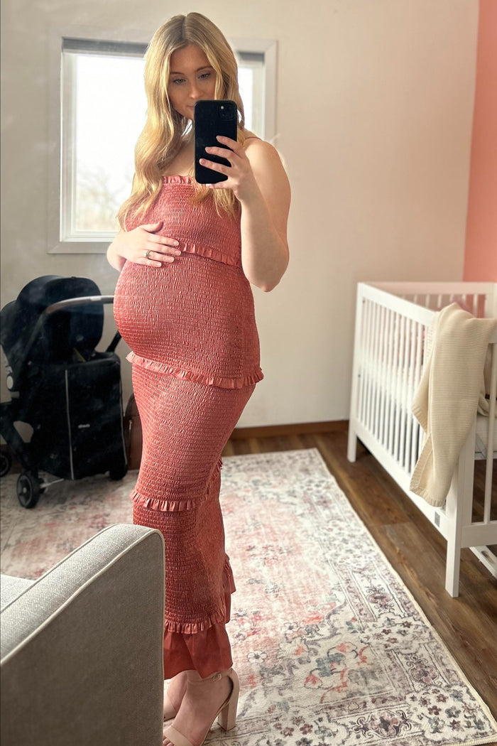 Pregnant woman in a smocked terracotta maternity dress taking a mirror selfie in a nursery with a white crib