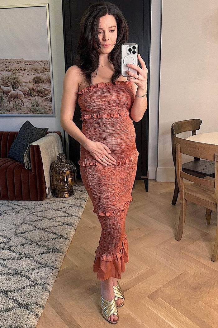 Pregnant woman in a terracotta smocked maternity dress taking a mirror selfie in a modern living room