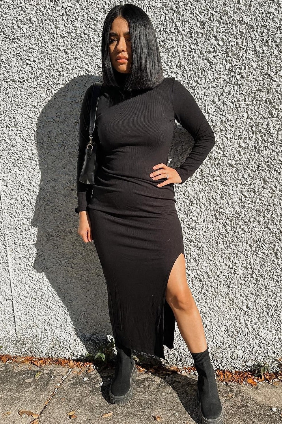 woman wearing a black long sleeve dress with a side slit and black ankle boots, standing against a textured wall
