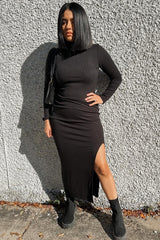 woman wearing a black long sleeve dress with a side slit and black ankle boots, standing against a textured wall