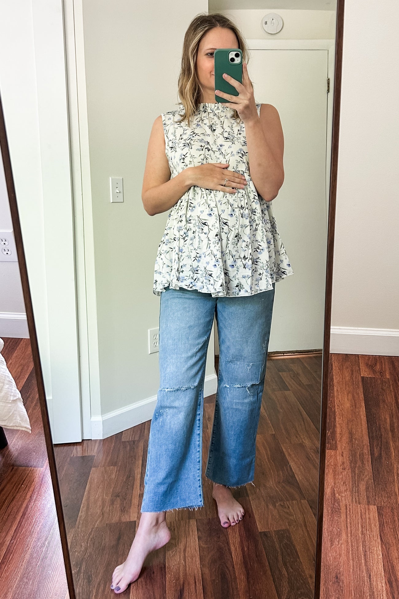 woman wearing a floral sleeveless maternity top and distressed blue jeans, holding her baby bump in front of a mirror