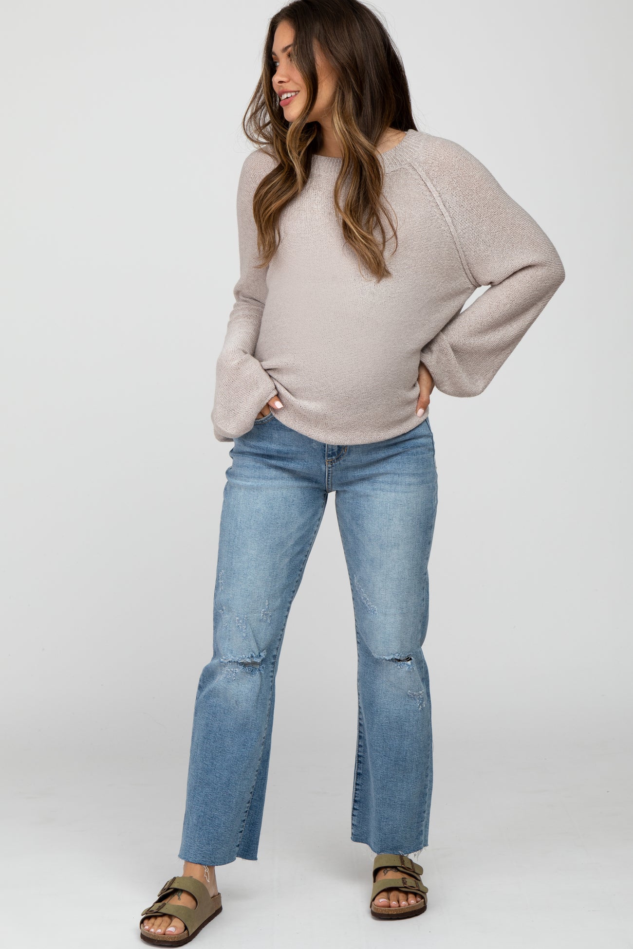 woman wearing a light beige maternity sweater with bell sleeves and distressed light blue jeans, showcasing her baby bump