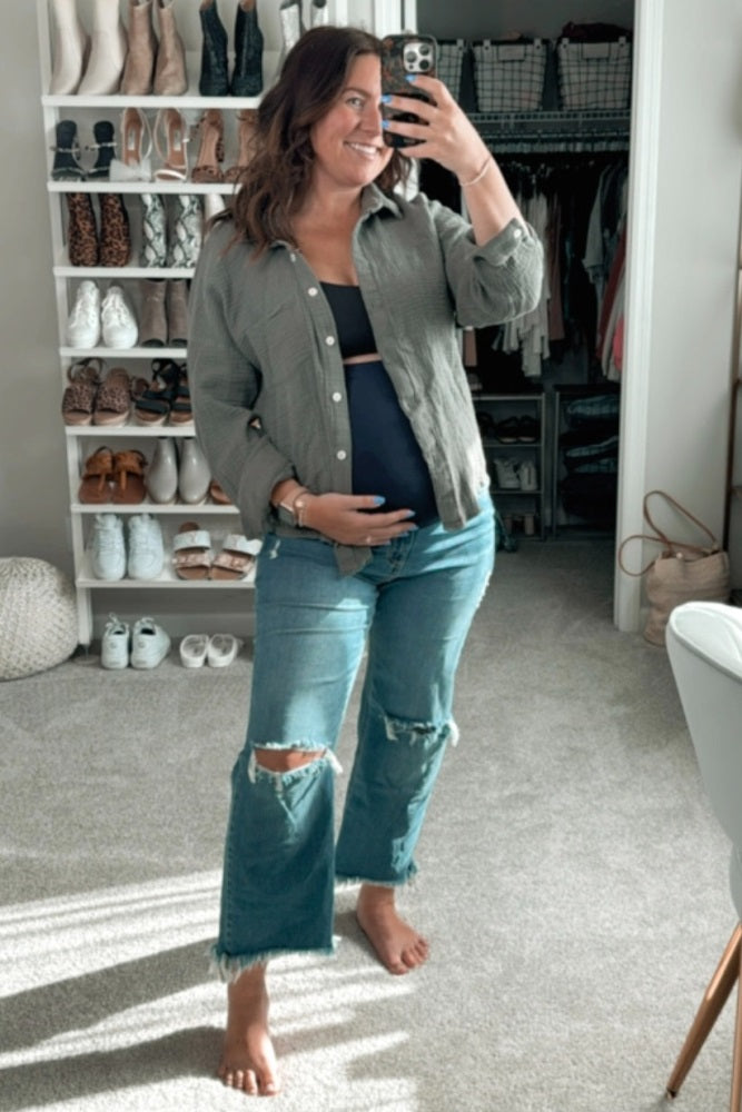 woman wearing a green button-up shirt and dark blue maternity top, showing her baby bump with distressed jeans