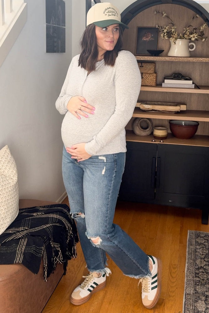 woman wearing a gray long sleeve maternity top and distressed jeans, holding her baby bump, styled with a cap and sneakers