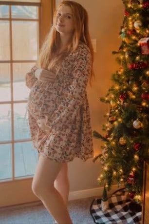 woman wearing a floral long sleeve maternity dress and holding her baby bump in front of a decorated Christmas tree