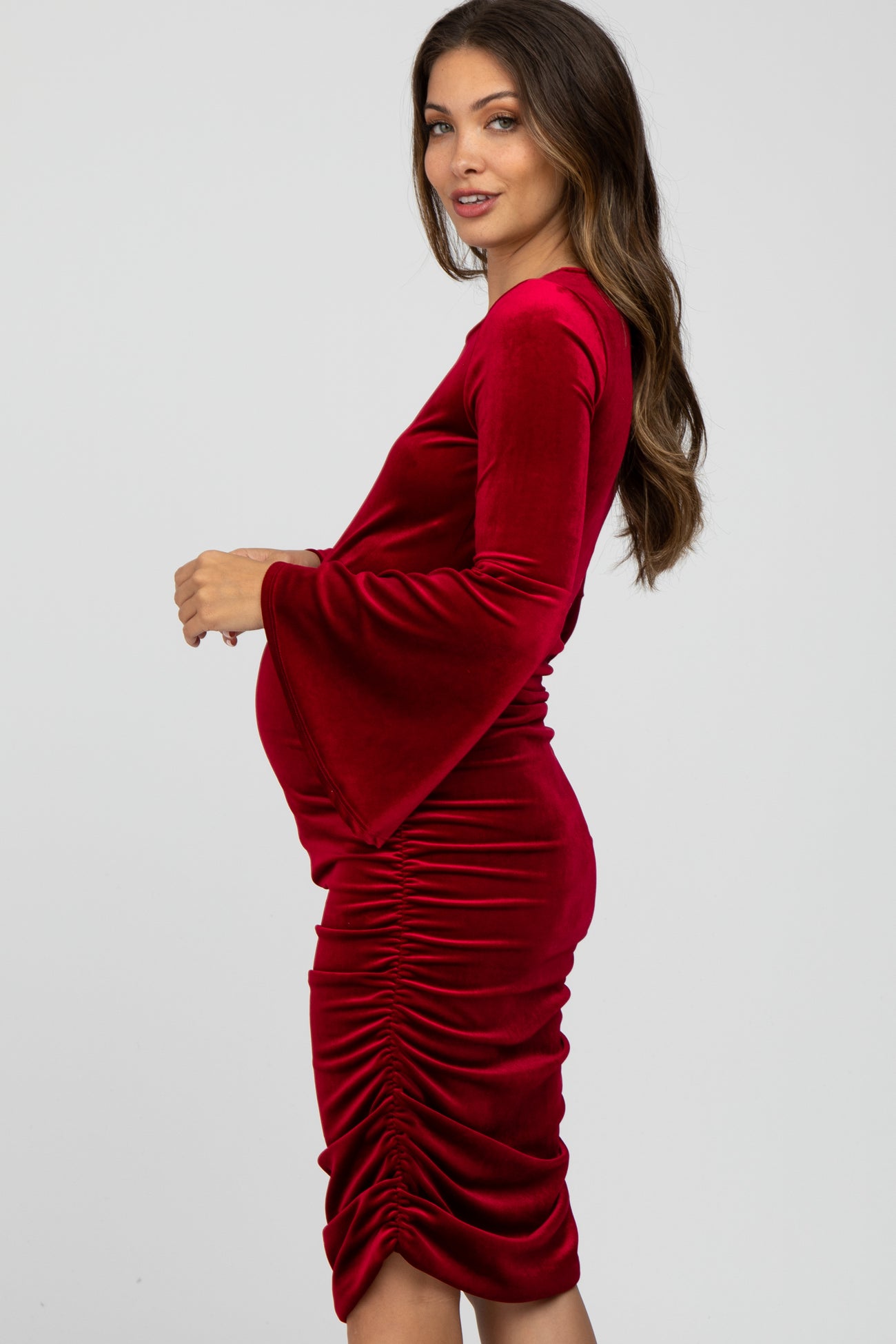 woman wearing a red velvet maternity dress with bell sleeves and side ruching, showcasing her baby bump