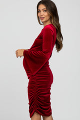 woman wearing a red velvet maternity dress with bell sleeves and side ruching, showcasing her baby bump