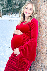 woman wearing a red velvet long sleeve maternity dress, showing her baby bump while leaning against a tree in snow