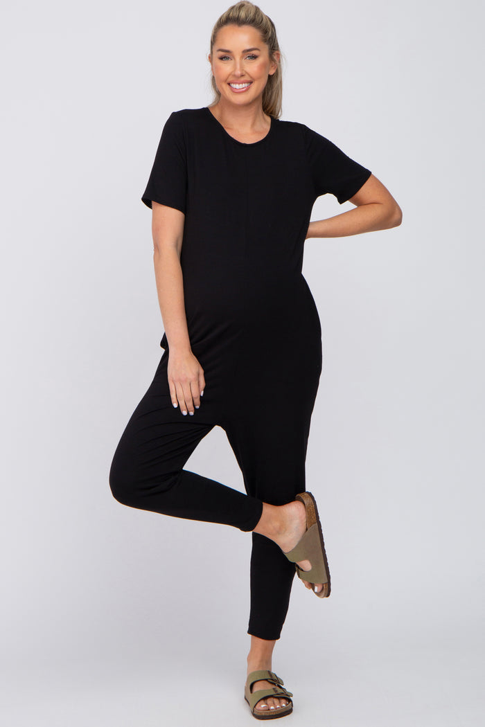 woman wearing a black short sleeve maternity jumpsuit and posing with one leg lifted, showcasing her baby bump