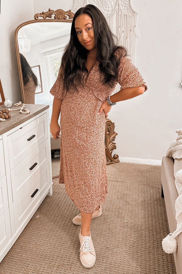 woman wearing a pink floral maternity dress and white sneakers, posing with one hand on her hip and a visible baby bump