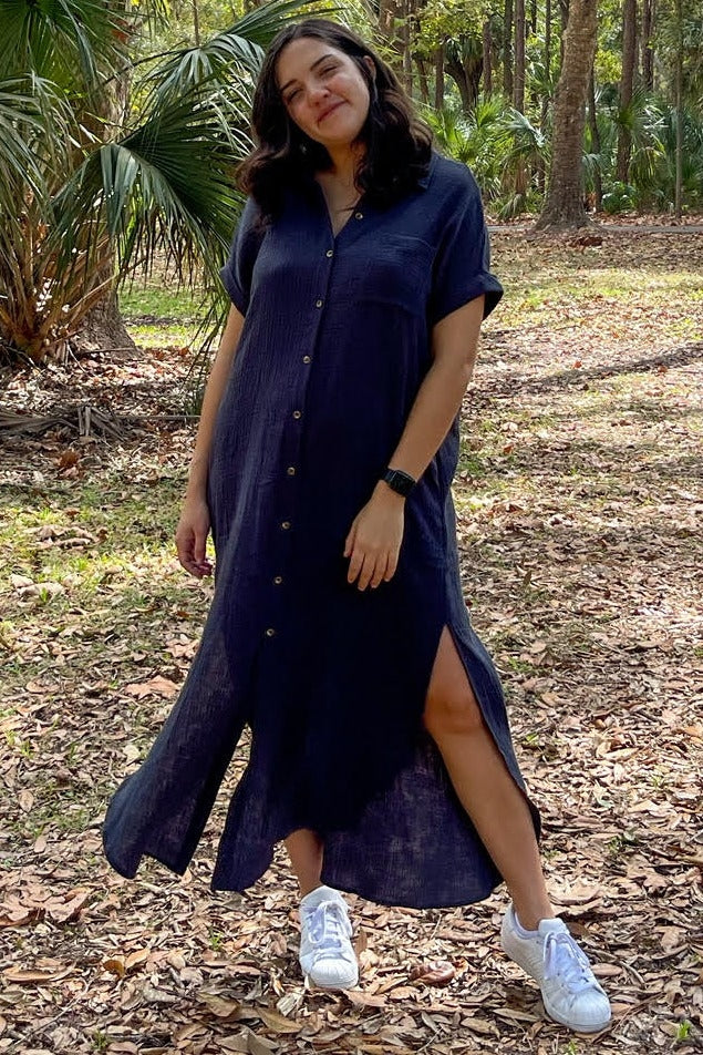 woman wearing a navy blue button-up maxi dress with side slits and white sneakers, posing outdoors in a forest setting
