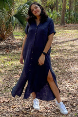 woman wearing a navy blue button-up maxi dress with side slits and white sneakers, posing outdoors in a forest setting