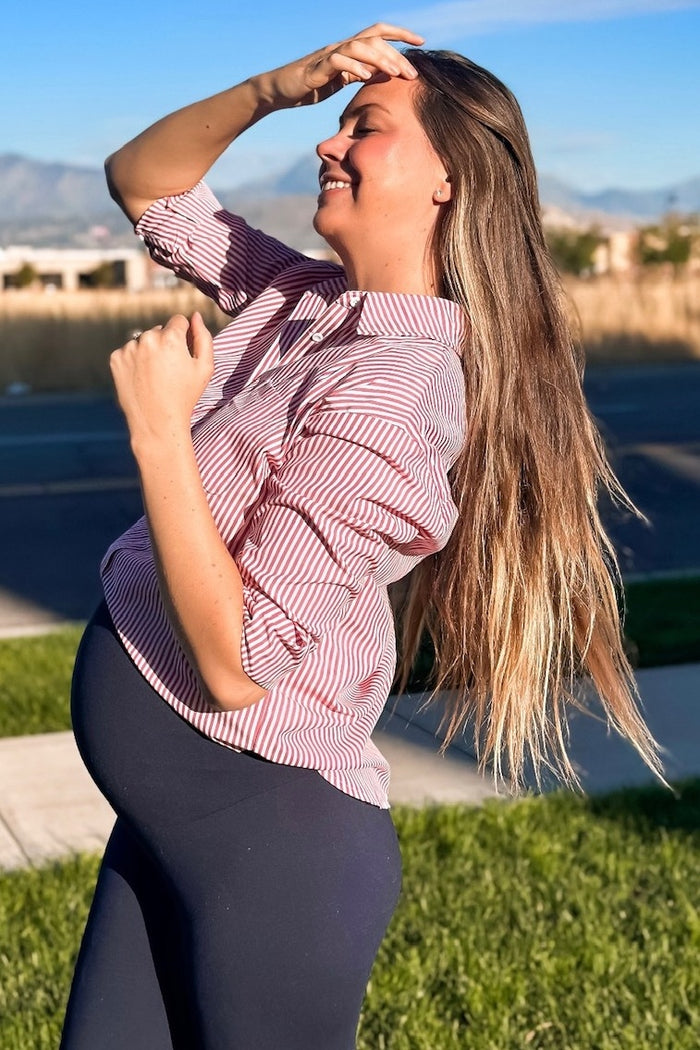smiling pregnant woman in red striped shirt and navy leggings standing outdoors in sunlight