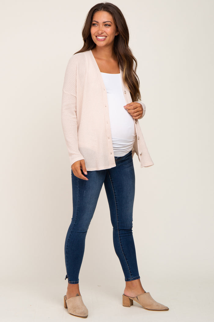 woman wearing a light pink maternity cardigan over a white top and blue maternity jeans, showing her baby bump