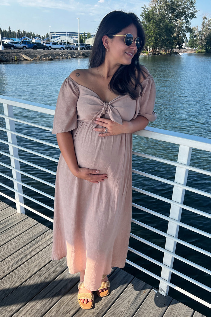 woman wearing a pink off-the-shoulder maternity dress with a tie detail, cradling her baby bump by the water