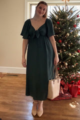 woman wearing a dark green midi dress with a front tie detail, standing near a decorated Christmas tree, holding a beige handbag