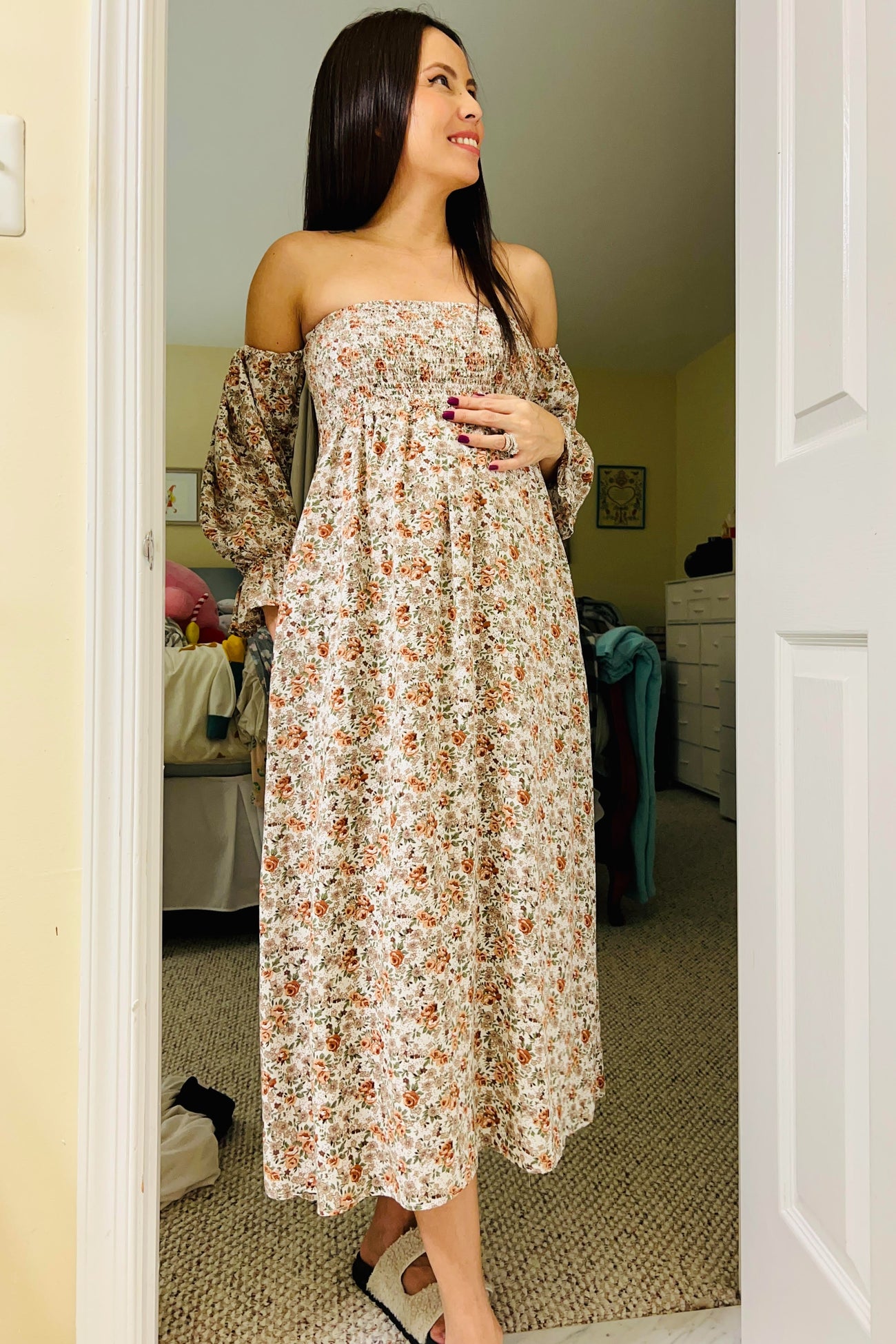woman wearing a floral off-the-shoulder maternity dress with a baby bump visible, standing in a bright room