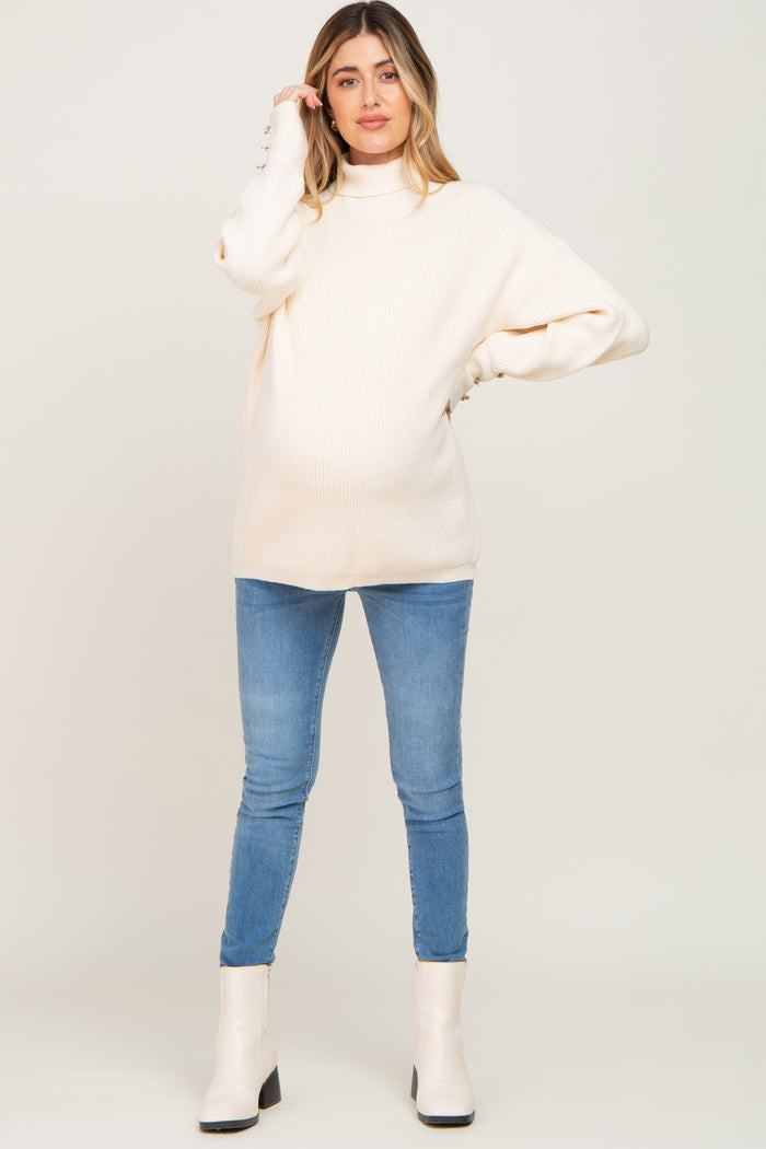 woman wearing a cream ribbed maternity sweater with a mock neck and blue skinny jeans, showing her baby bump