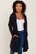 woman wearing a black long cardigan with a hood and side pockets, paired with a white tank top and blue jeans