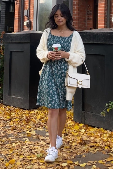 Woman in green floral dress and white cardigan holding coffee on autumn sidewalk with yellow leaves