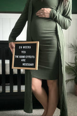 woman wearing a dark green ribbed maternity dress and long cardigan, holding a sign announcing 28 weeks pregnant with baby bump visible