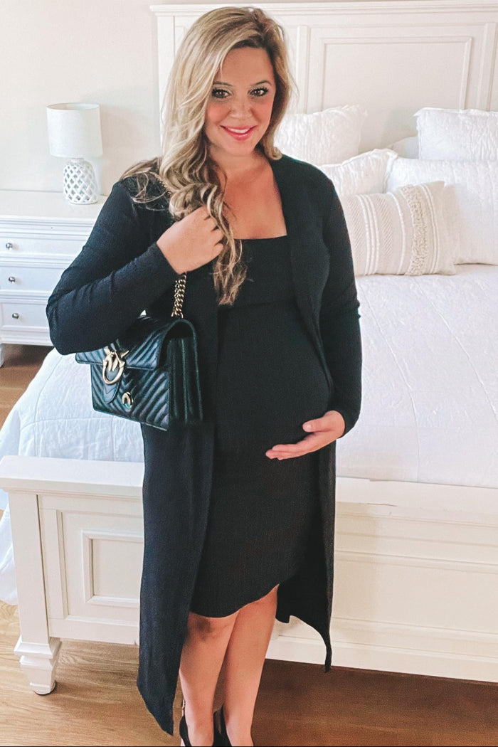 woman wearing a black maternity dress with a matching long cardigan and holding her baby bump