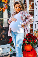 woman wearing a white tank top and a plaid shirt, showcasing her baby bump while standing near flowers and pumpkins