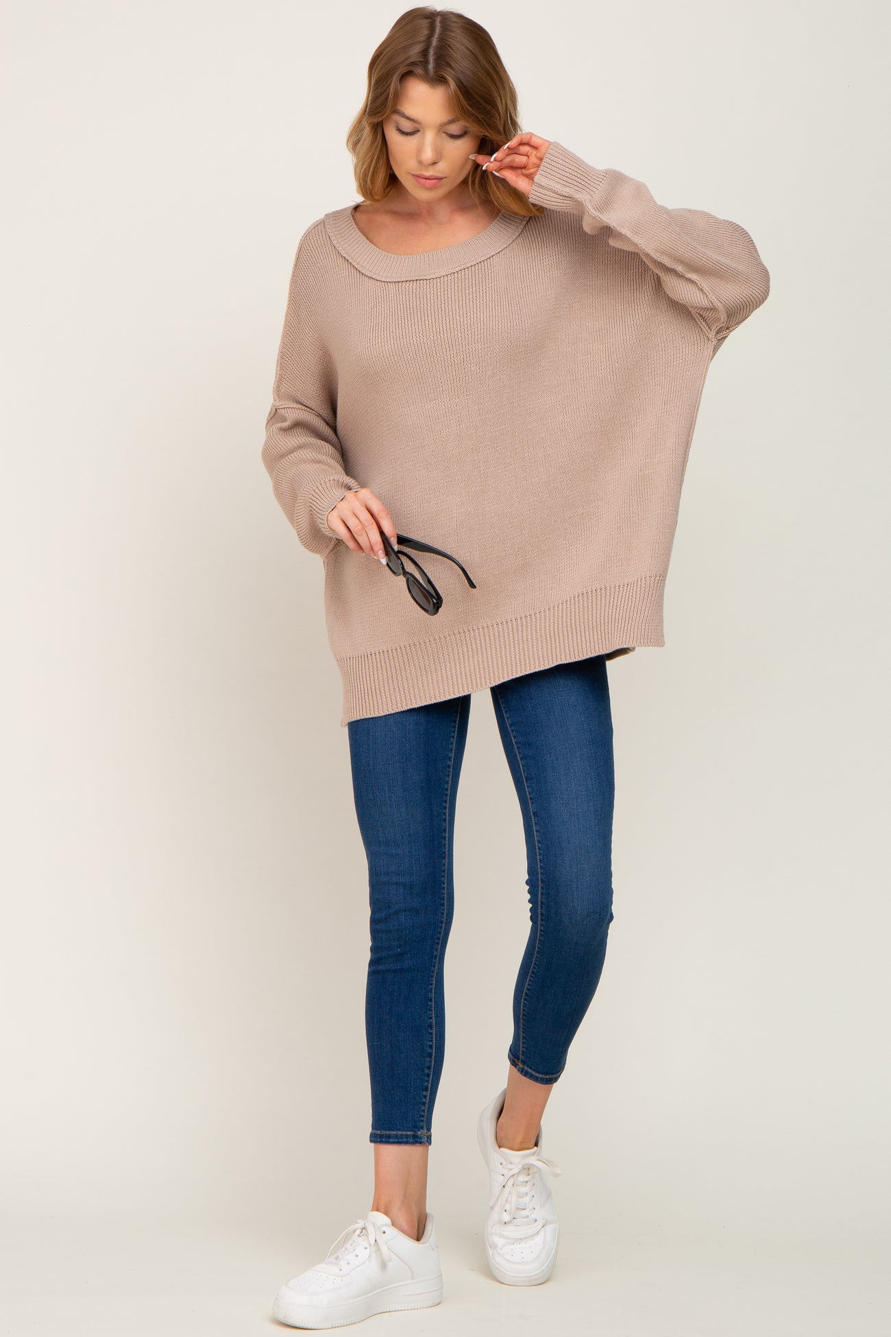 woman wearing a neutral beige oversized sweater and blue skinny jeans, holding sunglasses in her hand