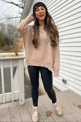 woman wearing a light pink oversized sweater, black leggings, and pink boots, smiling outdoors with a beanie on her head