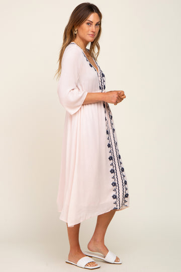 woman wearing a blush pink embroidered midi dress with bell sleeves and side slits, styled with white slide sandals