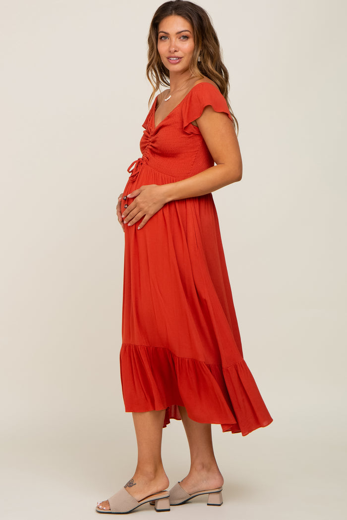 woman wearing a rust colored maternity dress with flutter sleeves and a ruffle hem, holding her baby bump