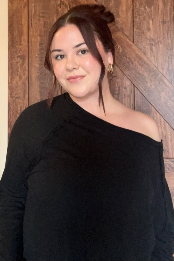 woman with dark hair in a bun wearing a black off-the-shoulder top in front of a wooden barn door