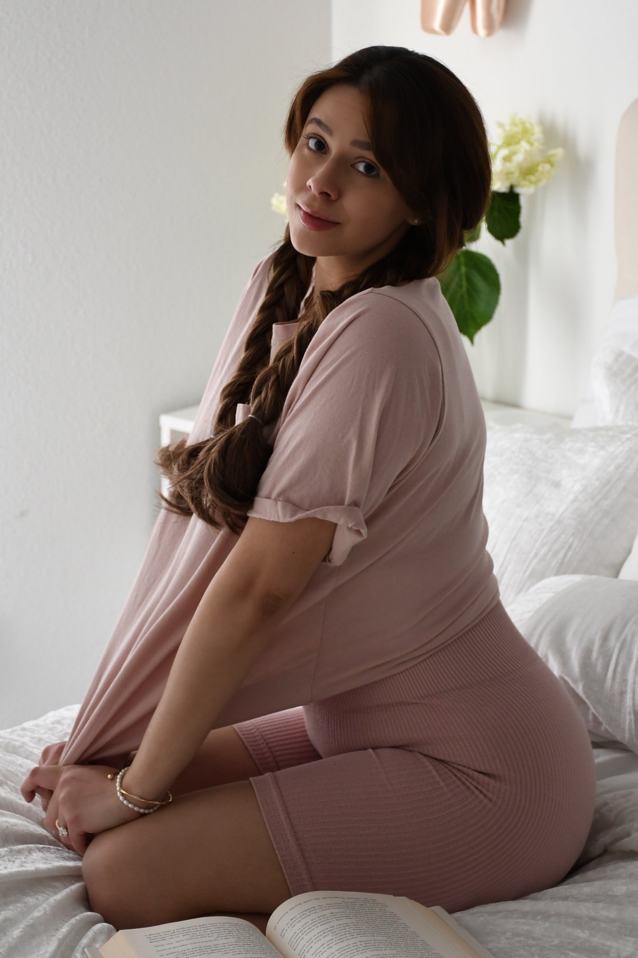 woman wearing a pink oversized top and matching ribbed shorts while sitting on a bed with a book and flowers nearby