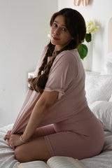 woman wearing a pink oversized top and matching ribbed shorts while sitting on a bed with a book and flowers nearby