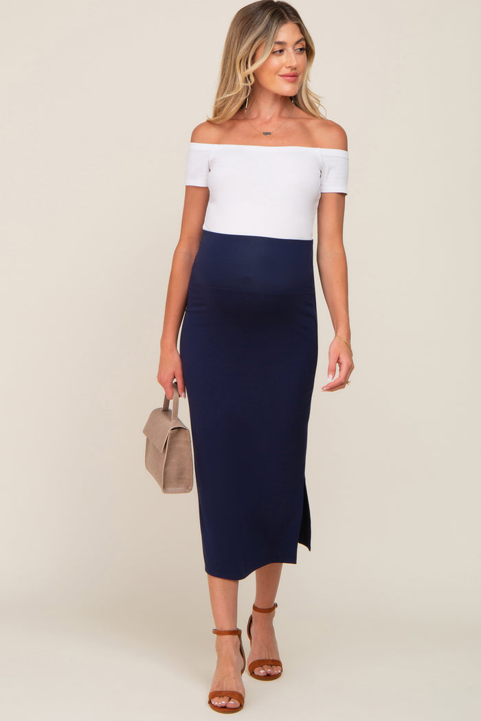 woman wearing a navy blue off-the-shoulder maternity dress with a white top and a side slit, holding a small handbag and showing her baby bump